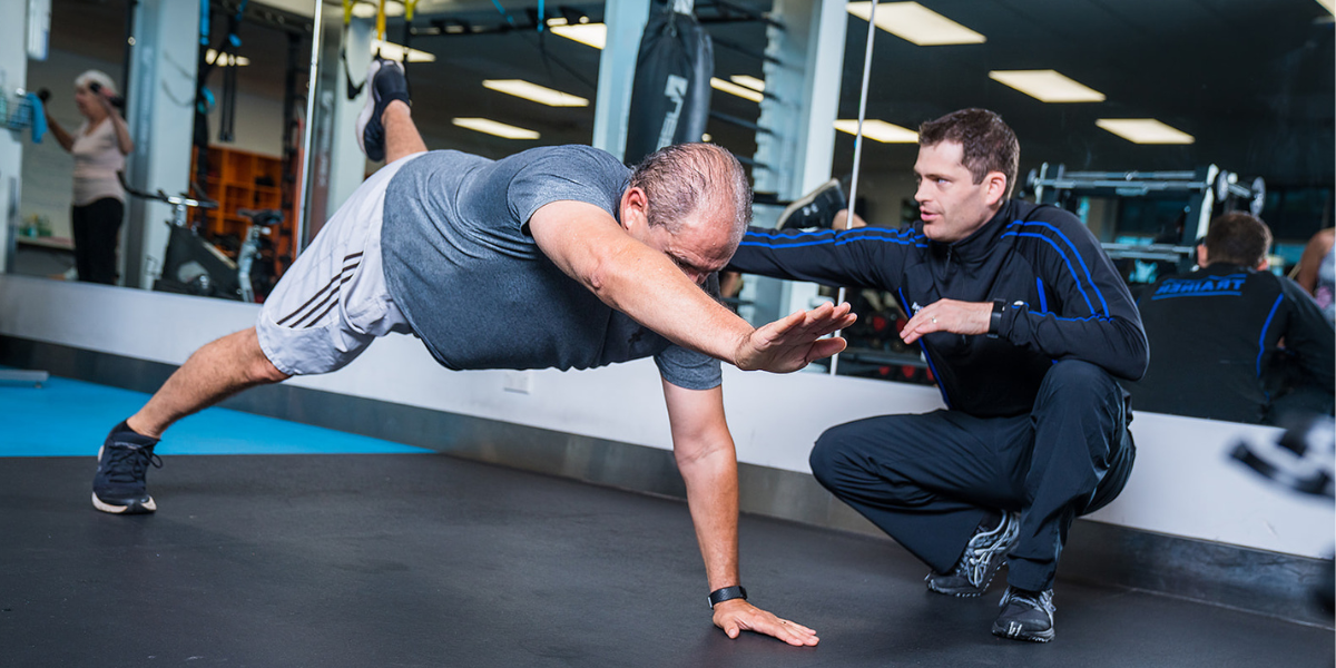 A male personal trainer assisting a man in blue with a core and leg workout at Christchurch Recreation and Sport Centre. 