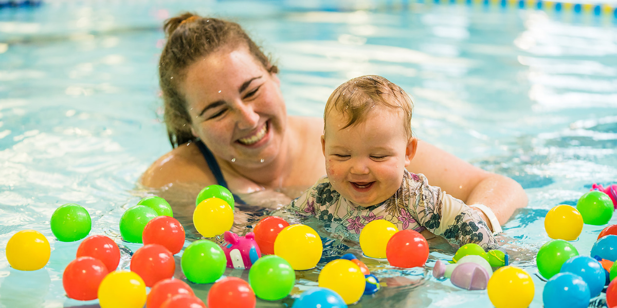 A smiling toddler playing with floating multicoloured balls while being held by a smiling woman at Christchurch Recreation and Sport pool. 