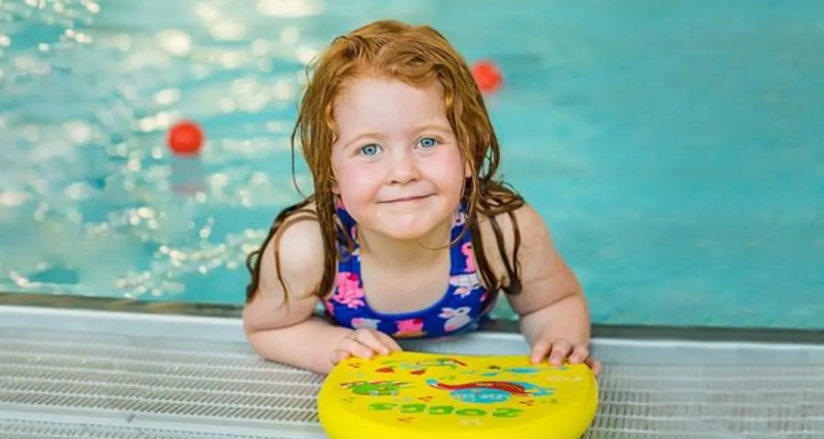 A smiling young girl holding onto a flotation board with swimming at Christchurch Recreation and Sport pool.