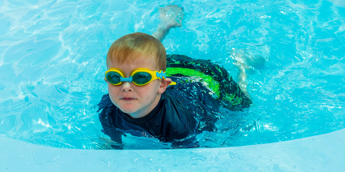 A little boy in yellow goggles and blue swimmers in the Waltham Summer toddler pool. 