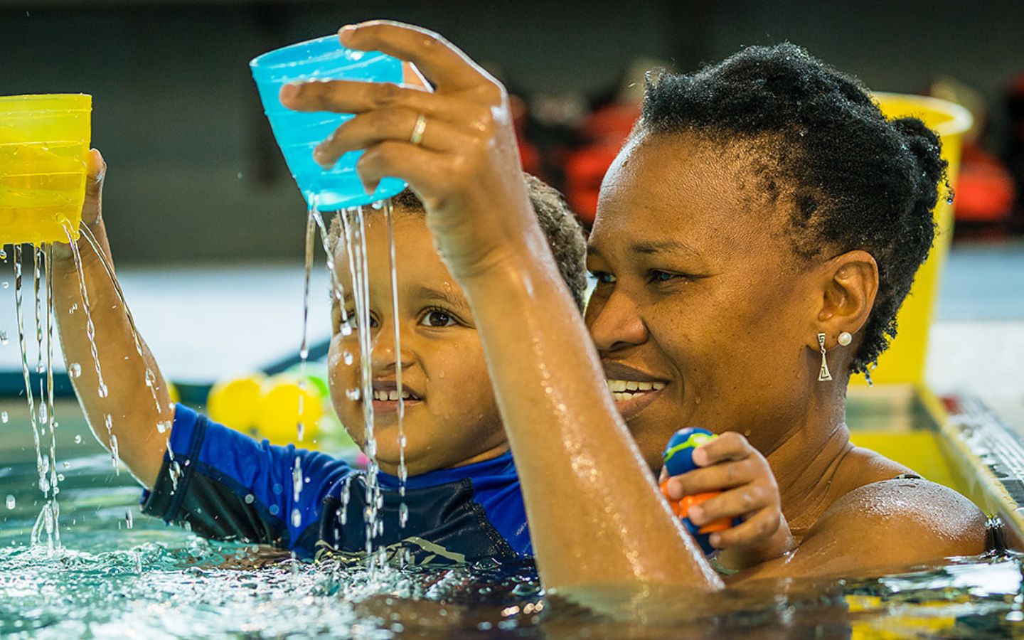 A little boy holding a yellow cup while being held by a smiling woman at Christchurch Recreation and Sport pool. 
