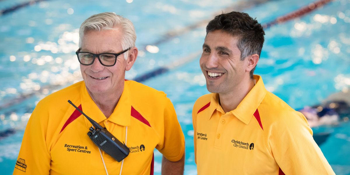Two smiling male lifeguards in yellow staff shirts in front of a Christchurch Recreation and Sport pool. 