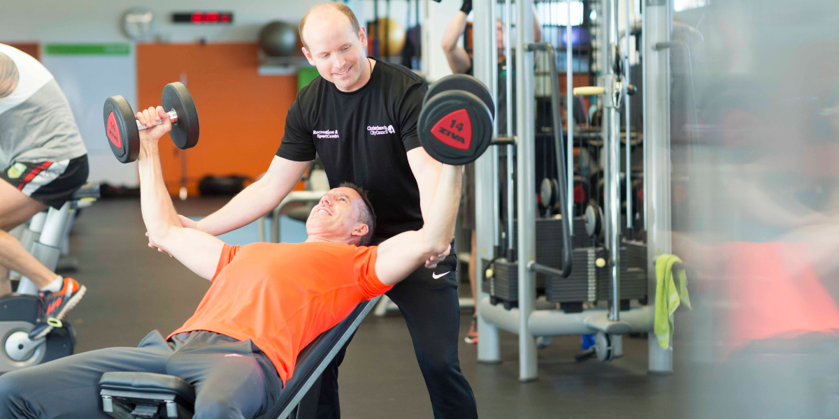 A male personal trainer in a black staff uniform assisting a male client in orange with a shoulder press at Christchurch Recreation and Sport Centre.