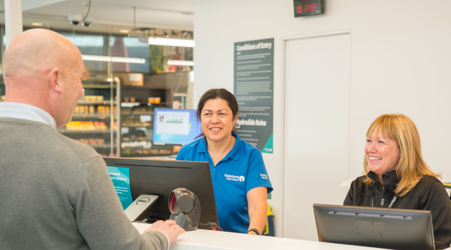 Two smiling women standing behind the counter at Jellie Park Rec Centre talking to a man in a grey sweater. 