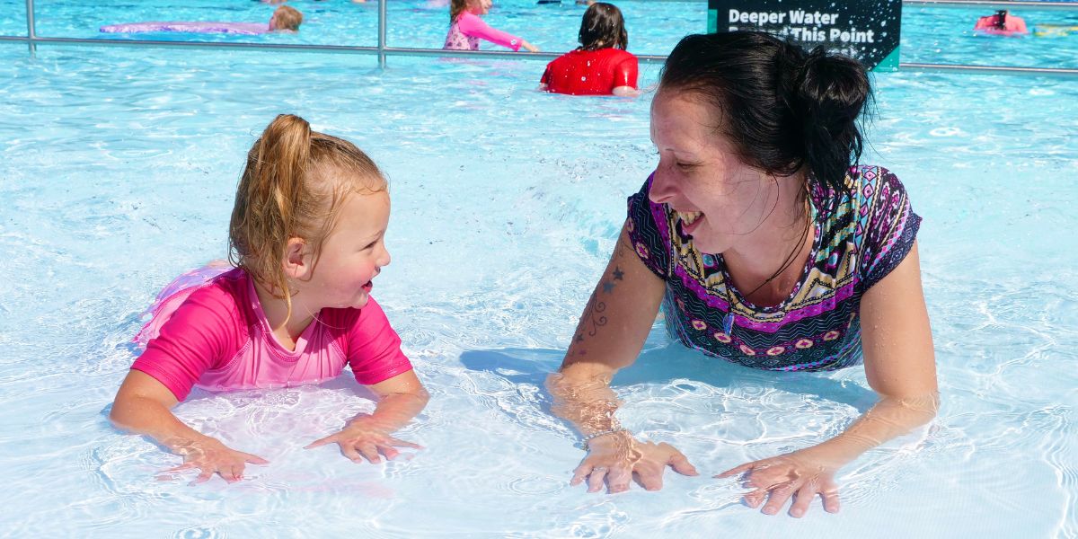 A child in pink and a smiling woman lay on their stomachs in the toddler pool at a Christchurch summer pool. 