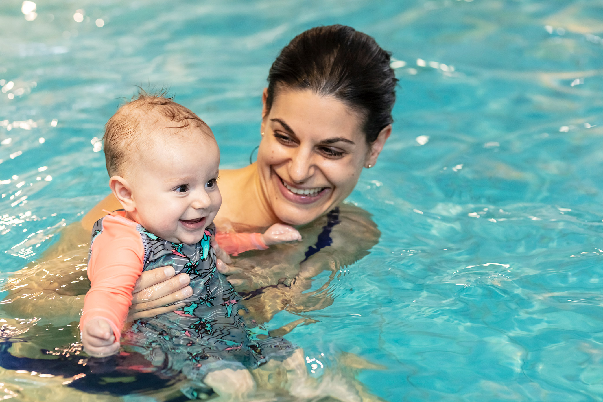 A smiling woman holding onto a baby while attending a Tiny Turtles swim class at Pioneer Recreation and Sport Centre. 